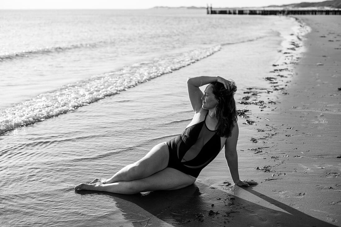 Vrouw in zwart badpak in zeemeermin pose in de branding van de zee tijdens een badpak shoot in Zeeland met prachtig tegenlicht op haar curves.