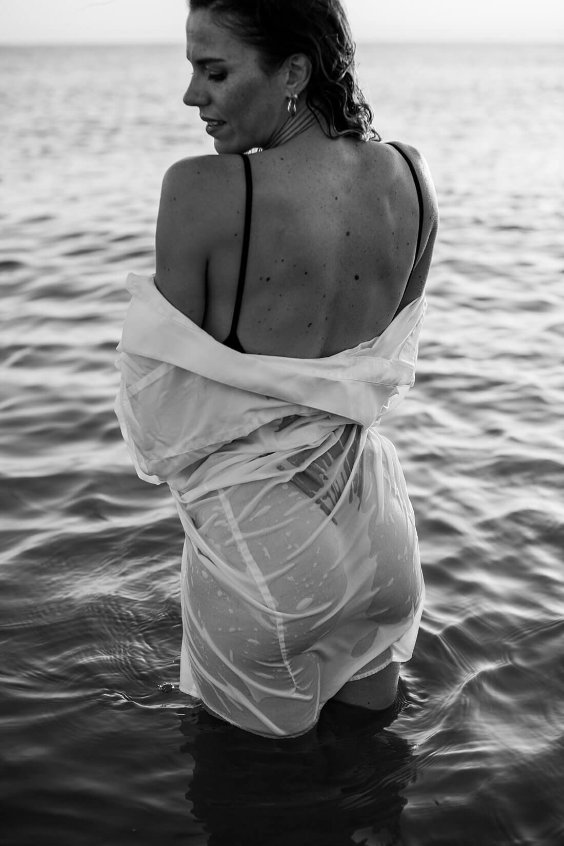 Zwart-wit semi close-up van een model in het water met een natte, doorschijnende oversized witte blouse tegen haar billen geplakt