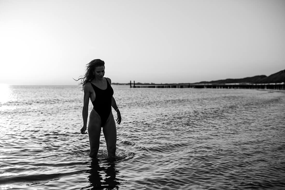 Zwart-wit silhouet van een vrouw in hooggesneden badpak lopend richting het strand met weerspiegeling van de zon