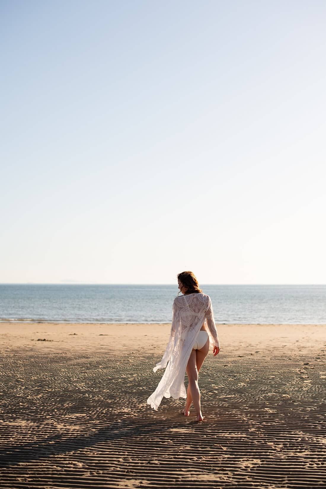 Model van achteren gefotografeerd in witte bikini en kanten kimono lopend richting de zee in Dishoek