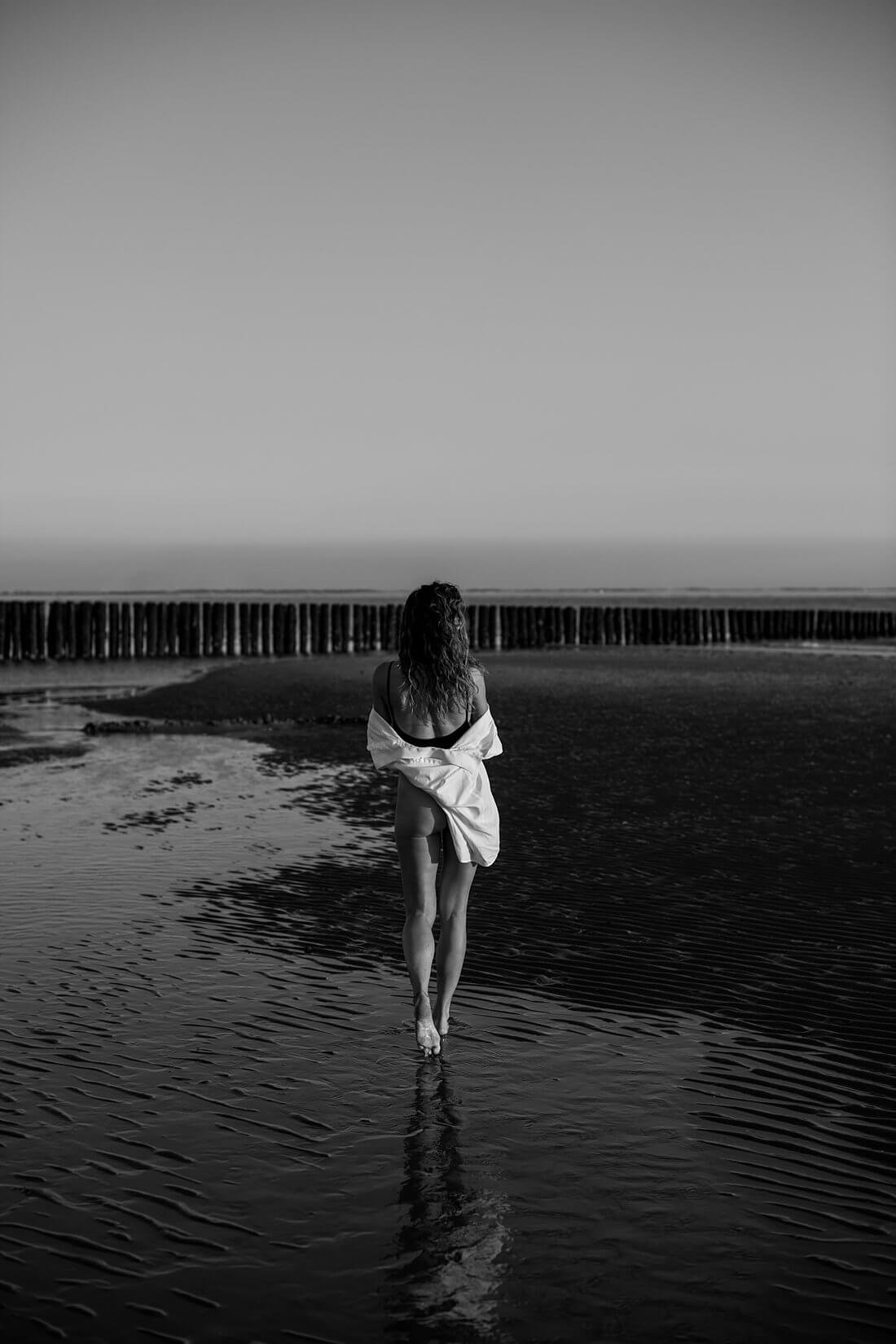 Model van achteren gefotografeerd lopend over een natte zandbank in zwart badpak met oversized witte blouse en weerspiegeling in het water