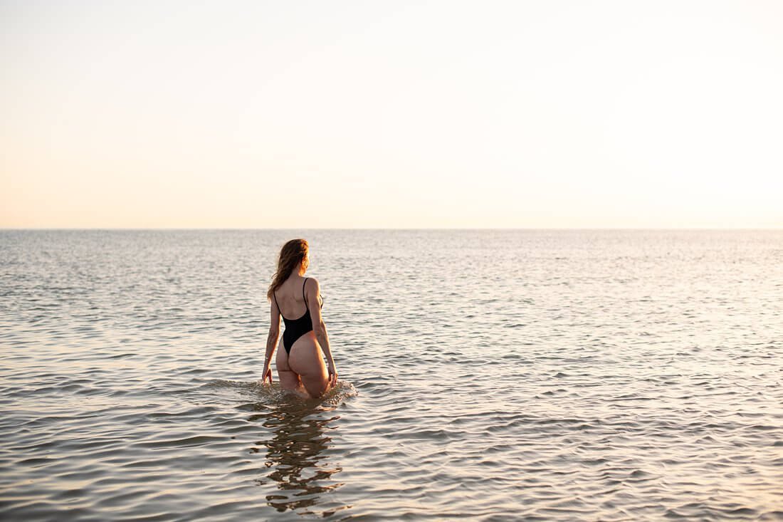 Dromerige kleurenfoto van de achterkant van een vrouw in zwart string badpak die tijdens golden hour de zee in loopt