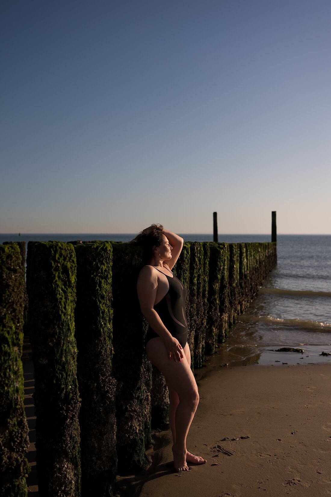 Vrouw in zwart badpak gefotografeerd van de zijkant met haar rug tegen de iconische strandpalen van Dishoek met contrastrijk zijlicht tijdens een boudoir fotoshoot.