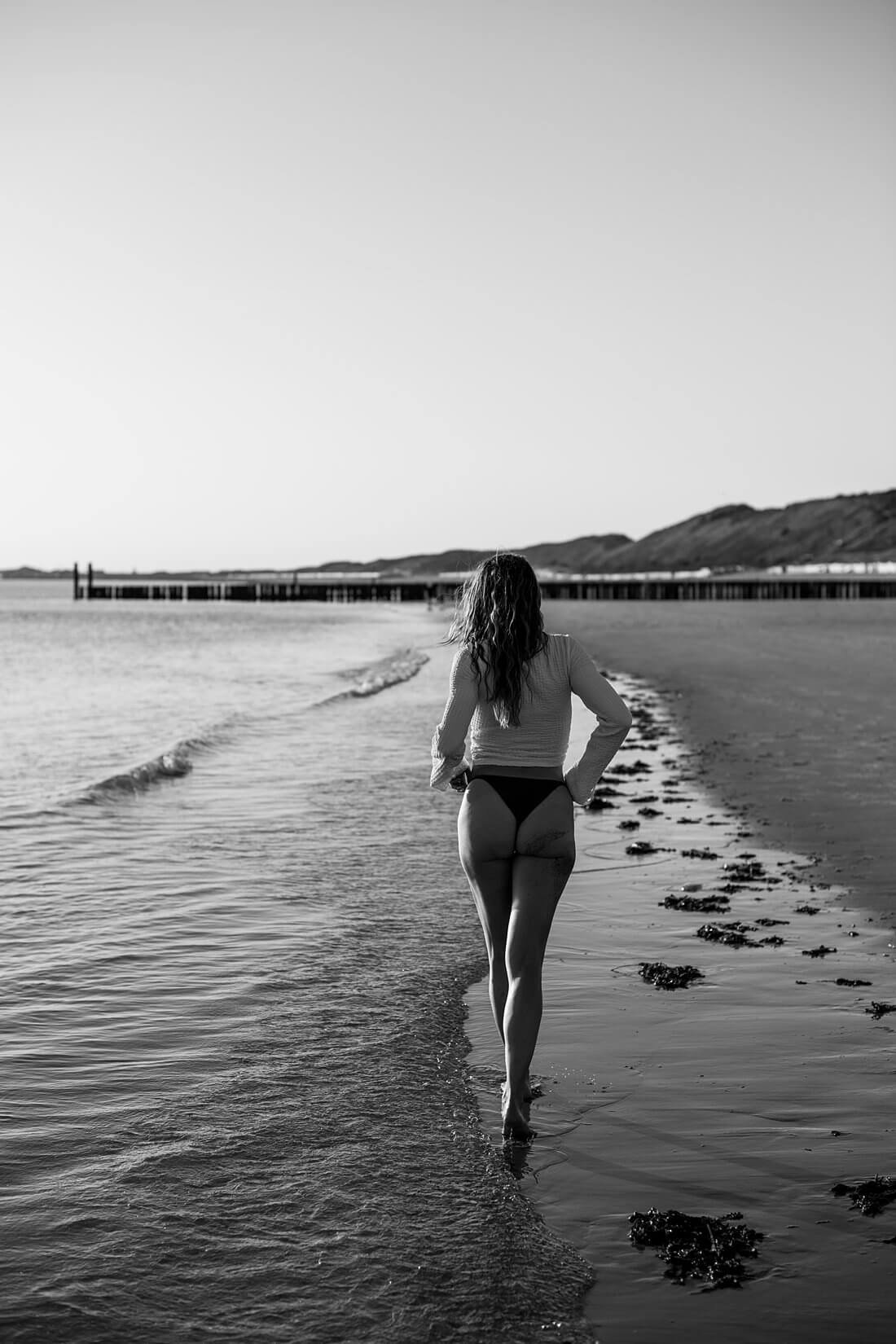 Vrouw loopt weg van de camera door de branding van de zee met tegenlicht tijdens een boudoir fotoshoot op het strand.