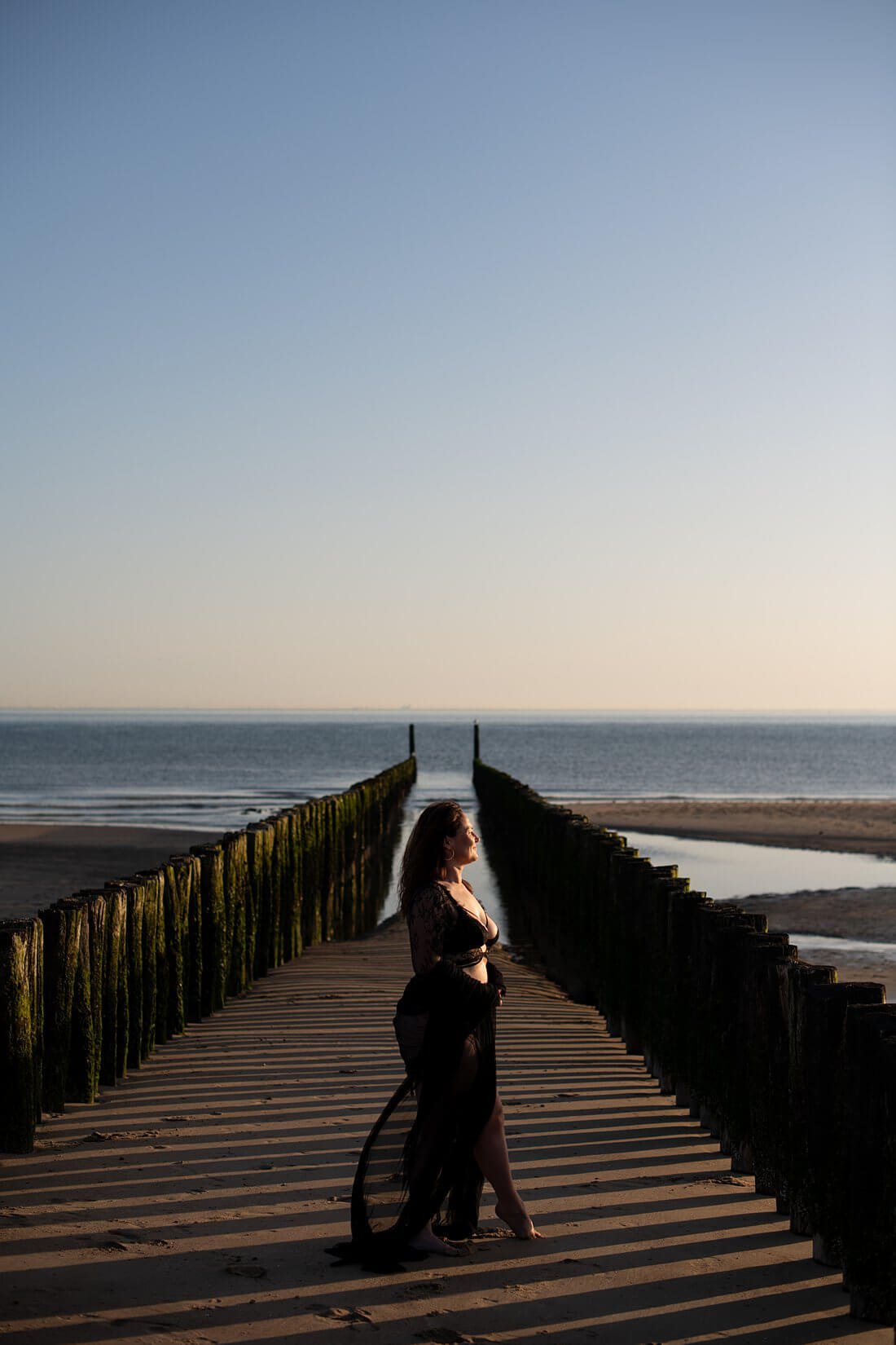 Vrouw staat zijwaarts in het midden tussen de strandpalen van Dishoek in Zeeland waarbij het licht prachtig op haar gezicht valt tijdens een boudoir fotoshoot.