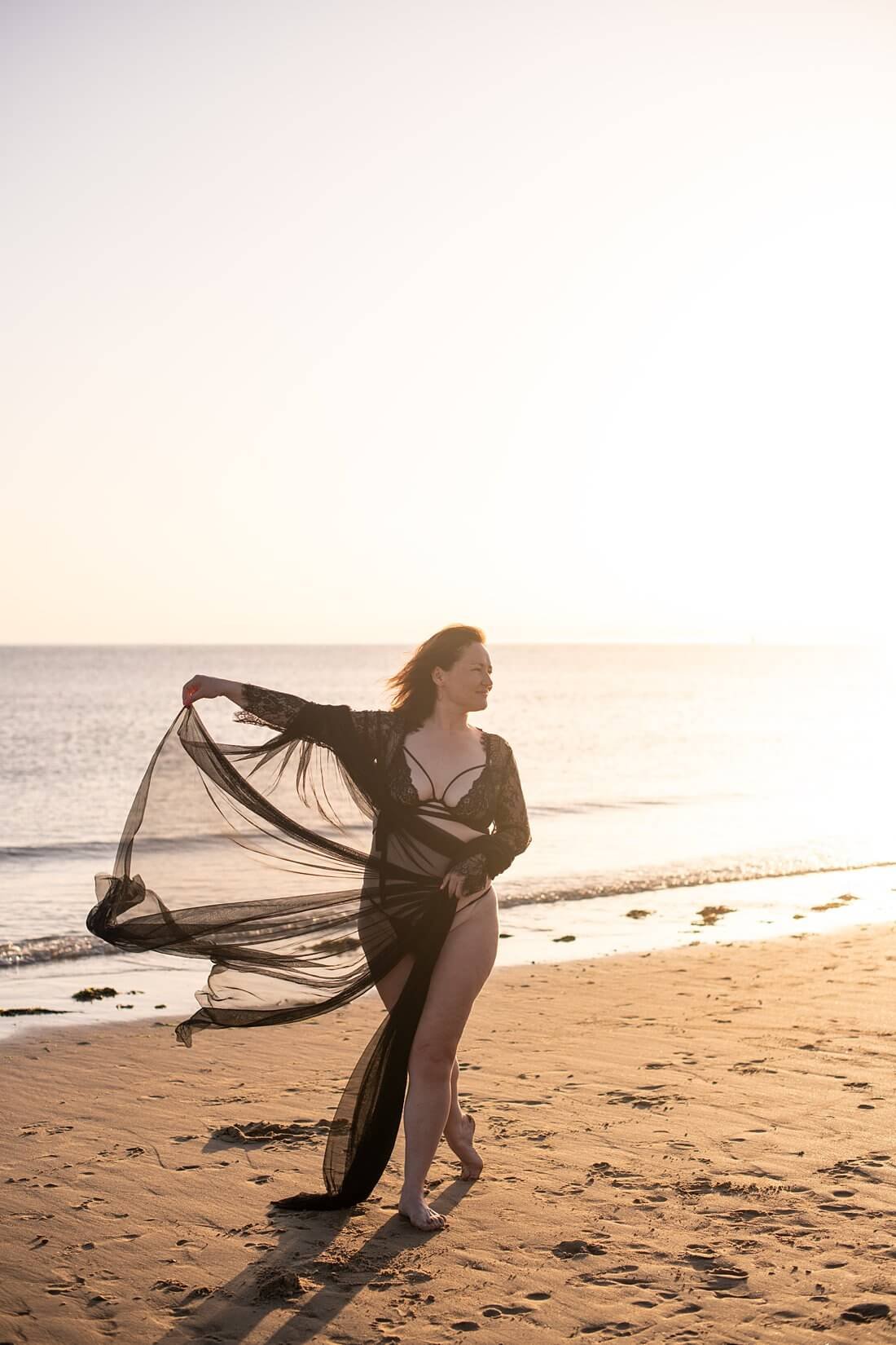 Dromerig beeld in kleur van een vrouw die een mesh doek vasthoudt in de wind met prachtig tegenlicht tijdens een badpak shoot in Zeeland.