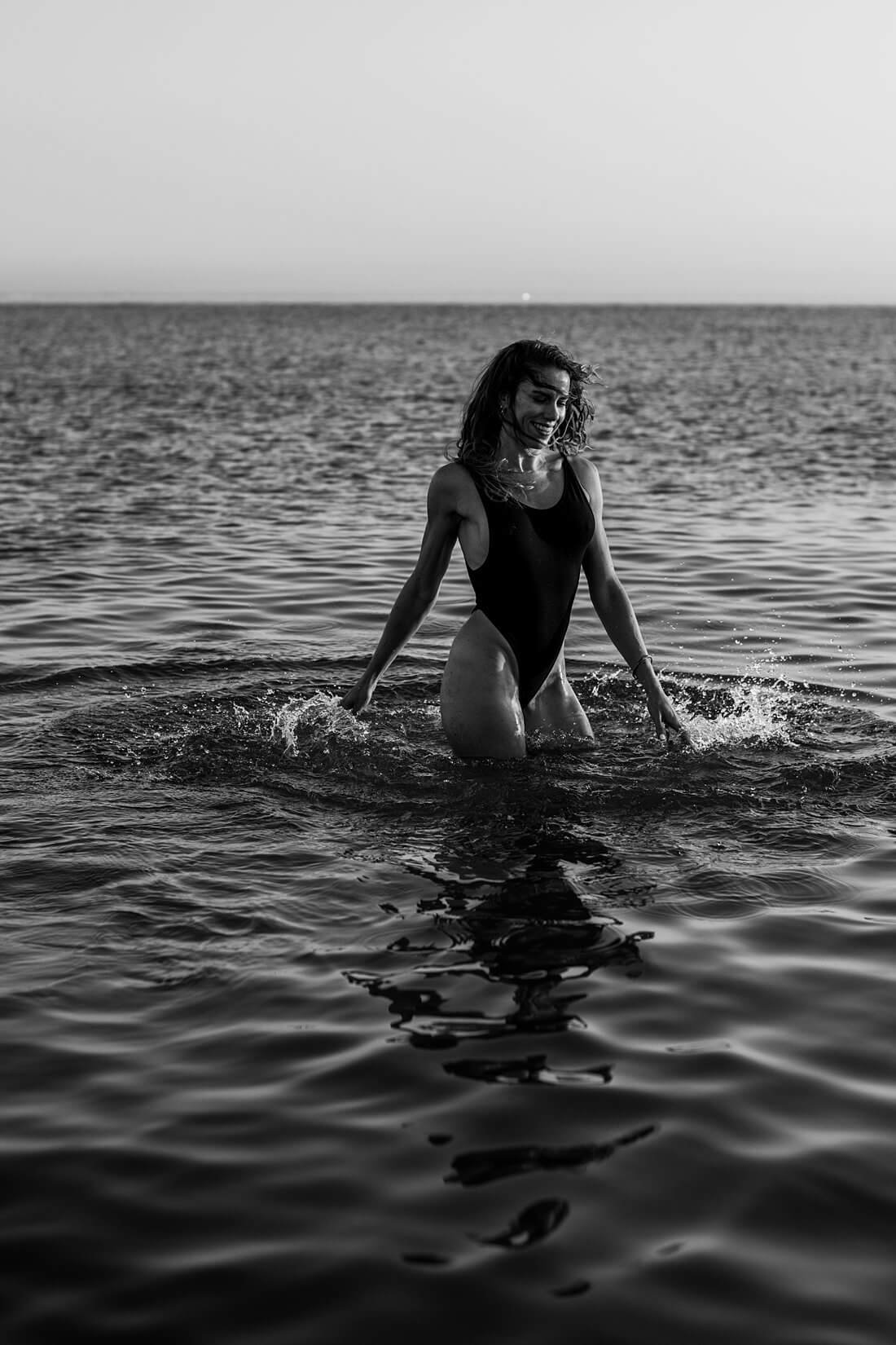 Zwart-wit actiefoto van een ronddraaiende, lachende vrouw in de zee met opspattend water