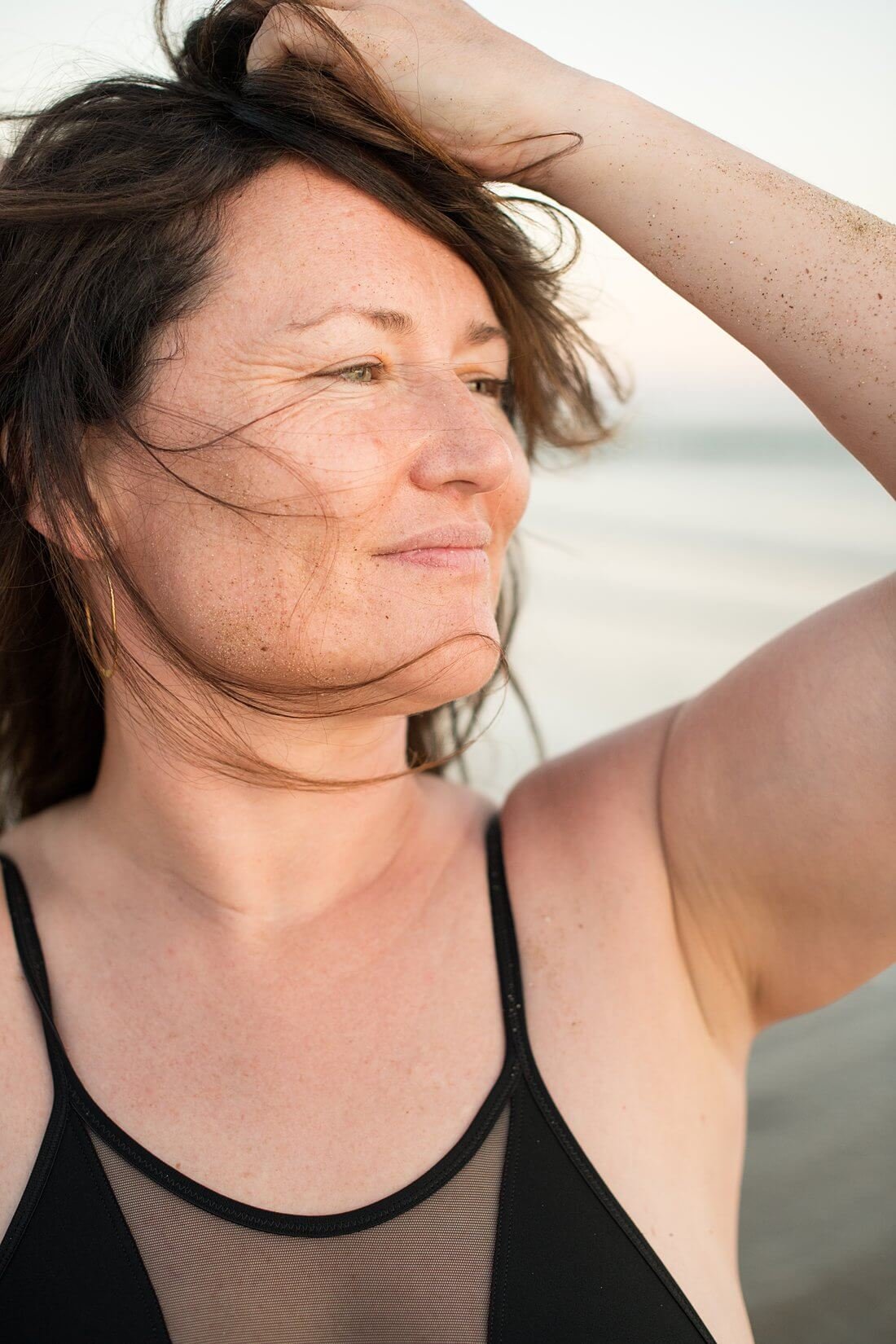 Portret in kleur van een vrouw met haar hand in haar haren die in het gouden zonlicht over de zee wegkijkt tijdens een strand shoot.