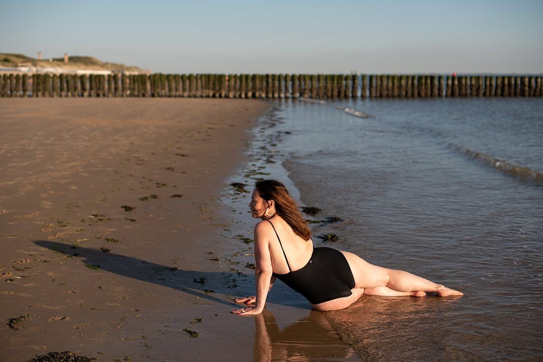 Plussize model in zwart badpak poseert van de achterkant in de branding tijdens een boudoir fotoshoot op het strand.