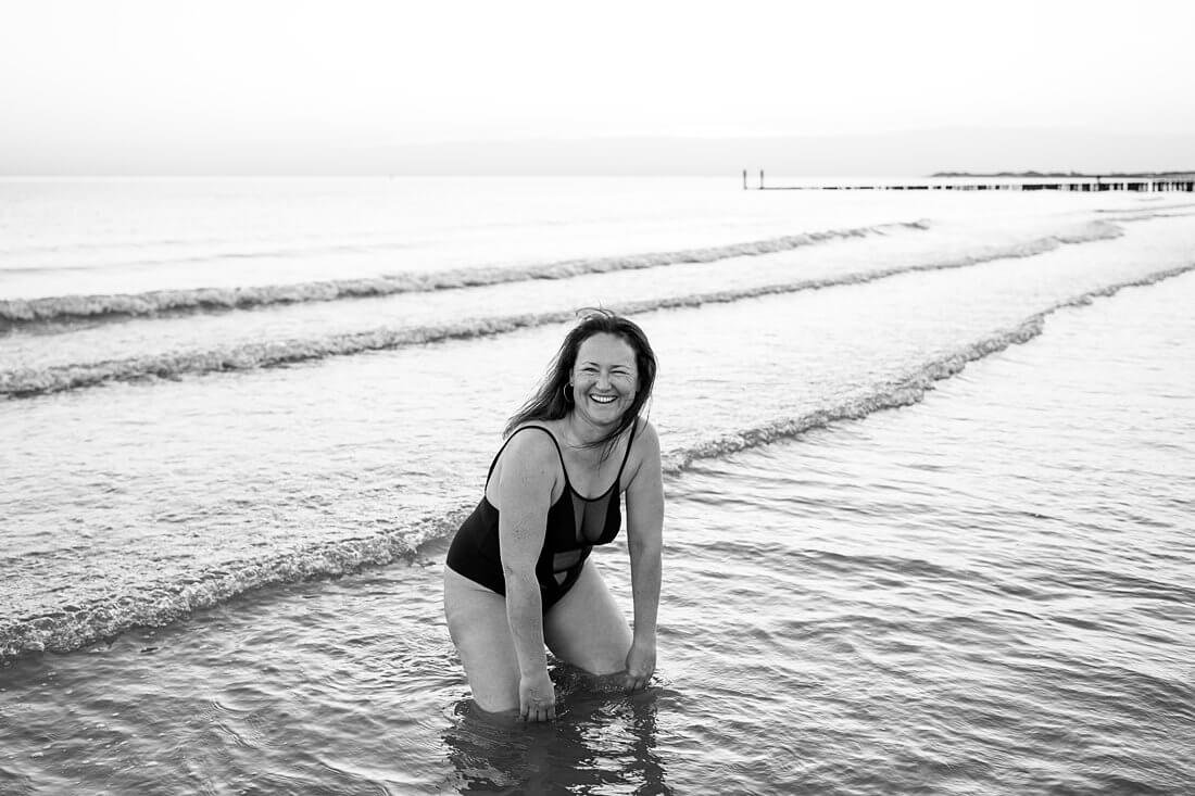 Speels zwart-wit beeld van een vrouw die op haar knieën in de branding van de zee zit en lachend in de camera kijkt tijdens een fotoshoot op het strand.