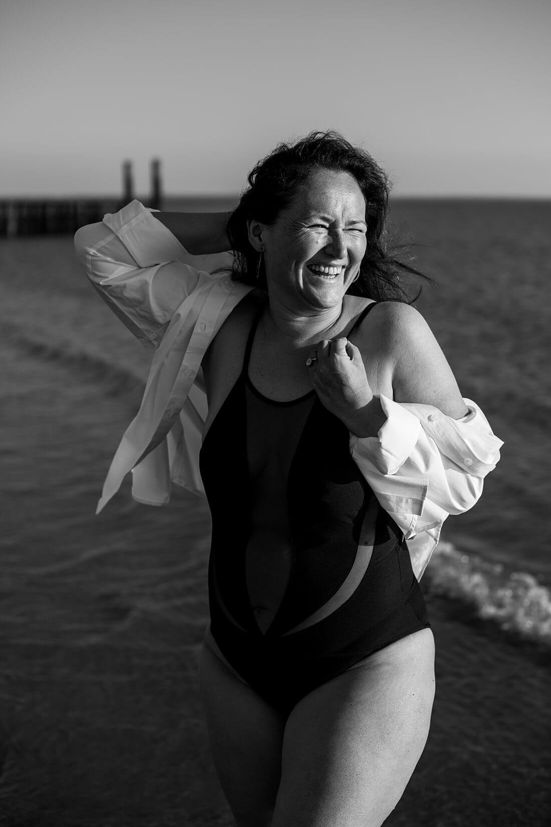 Semi close-up van een lachende vrouw met een oversized witte blouse en zwart badpak in de branding van de zee tijdens een boudoir fotoshoot op het strand.