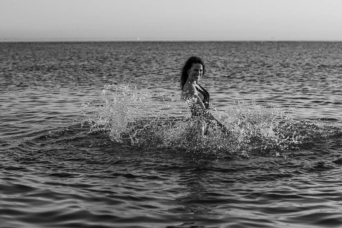 Lachende vrouw in zwart badpak springt rond in het opspattende zeewater tijdens een fotosessie op het strand.