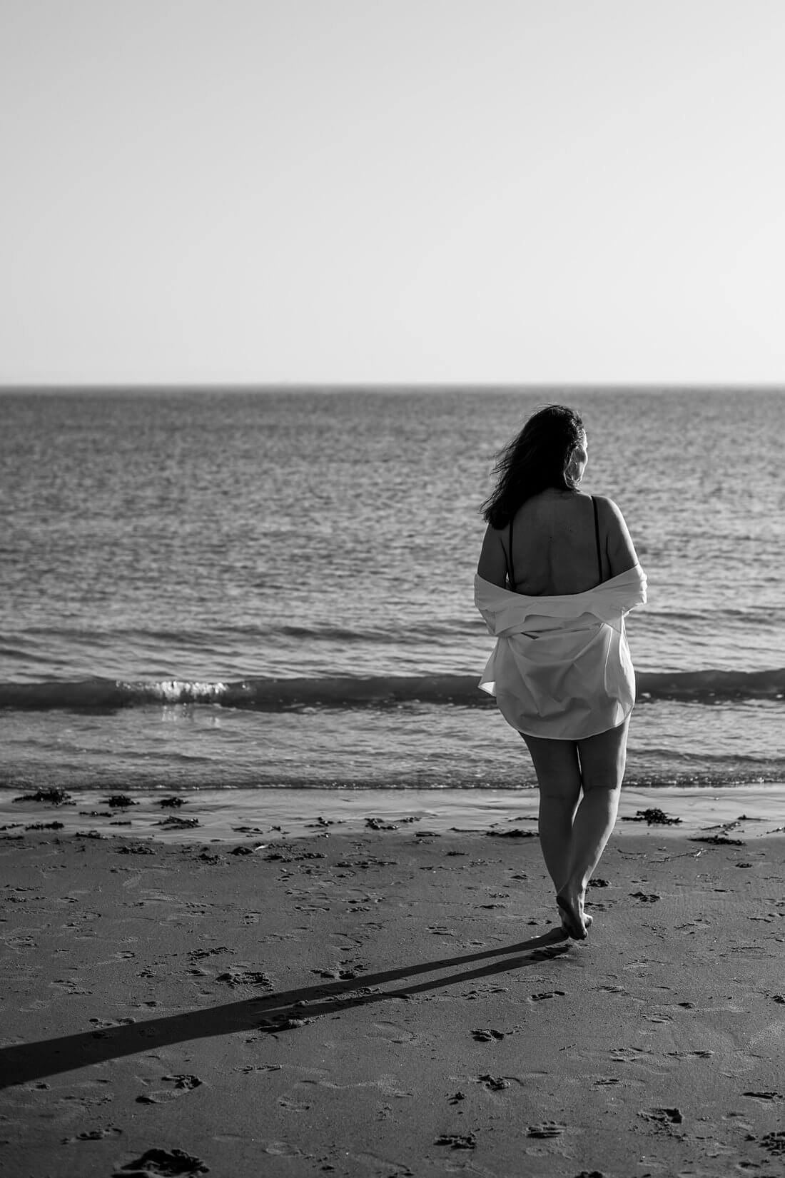 Achterkant van een vrouw in een badpak met lage rug en afgezakte witte blouse die over het strand naar de zee loopt tijdens een strand shoot in Zeeland.