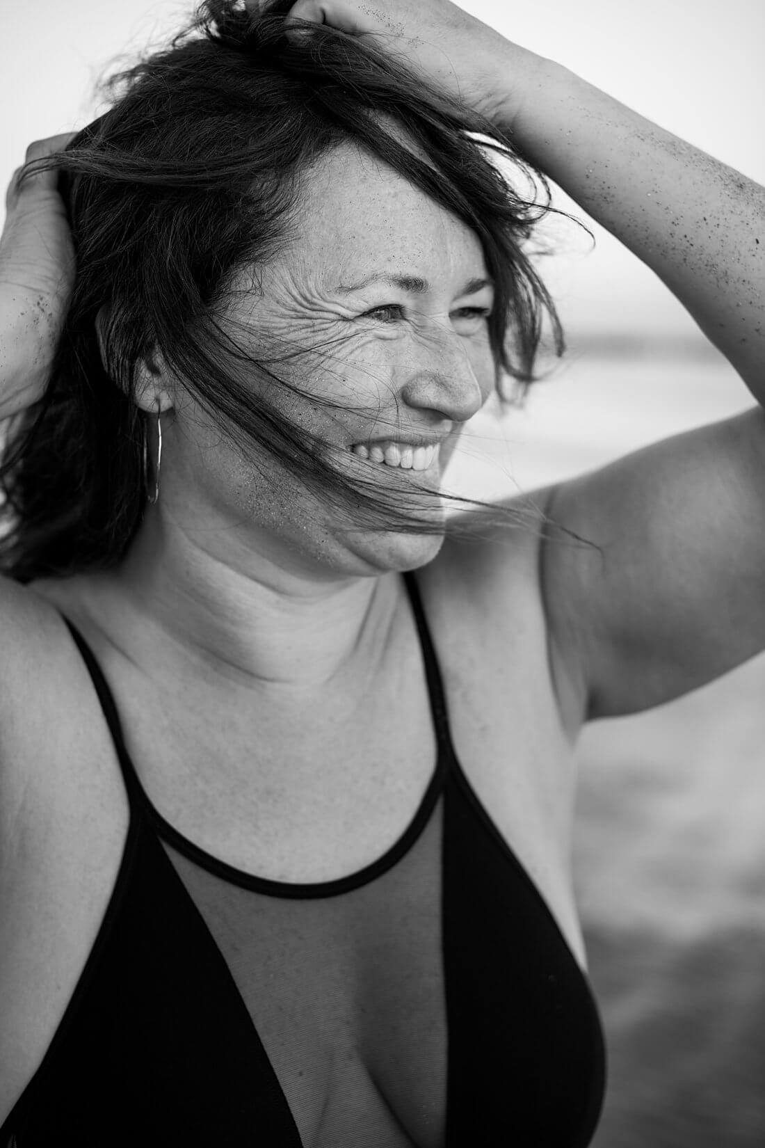 Prachtig zwart-wit portret van een lachende vrouw die wegkijkt terwijl de wind haar haren half voor haar gezicht blaast tijdens een strand shoot.