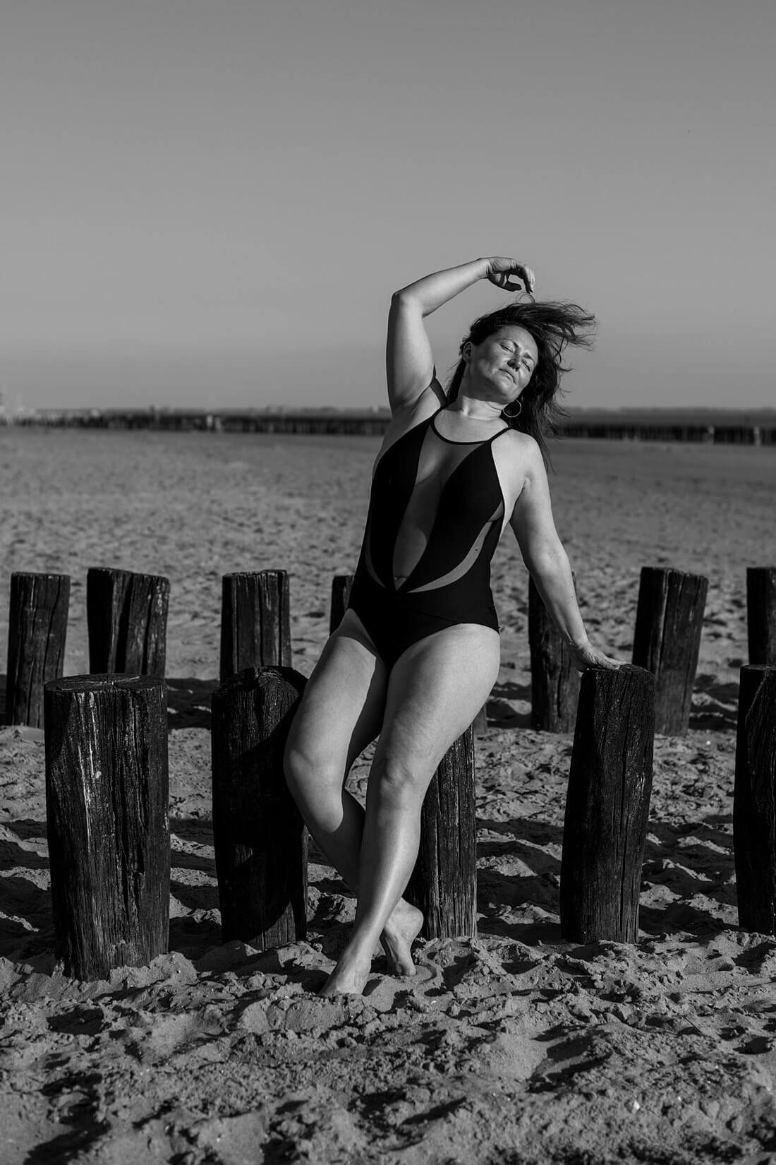 Zwart-wit boudoir strand shoot in Dishoek waarbij een vrouw met curves in zwart badpak languit gestrekt op de strandpalen zit en met haar hand door haar waaiende haren speelt.