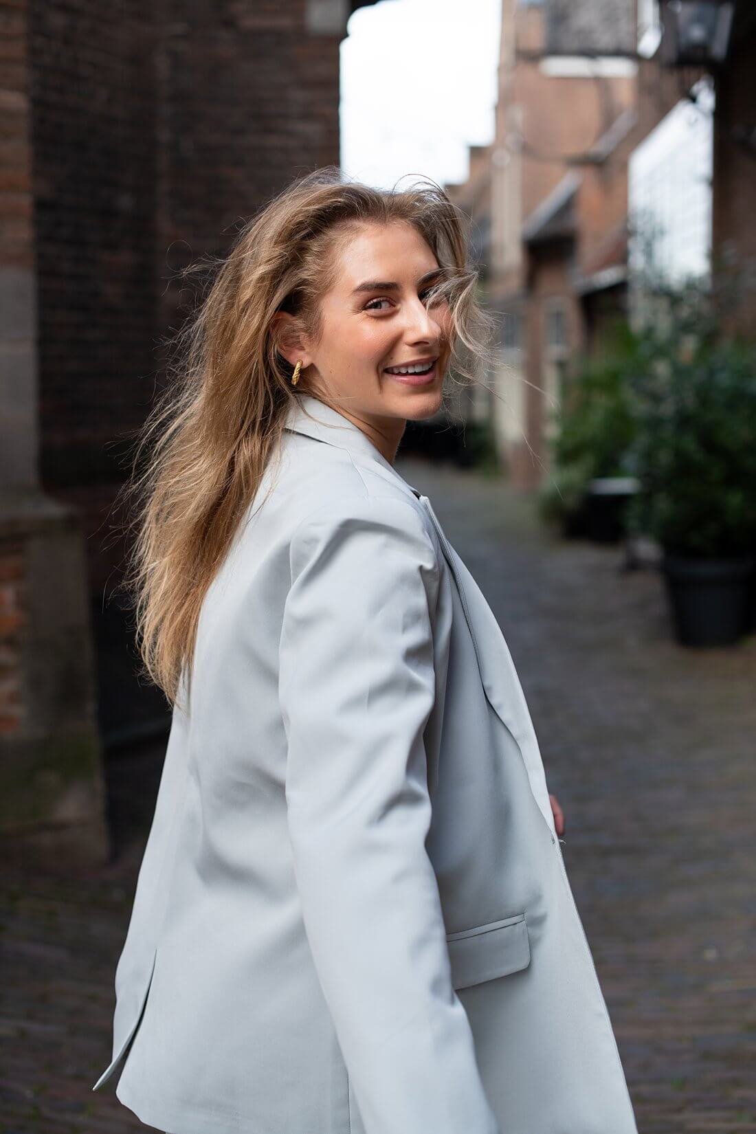 Sarah kijkt lachend over haar schouder in een lichtgrijs pak in Nijmegen.