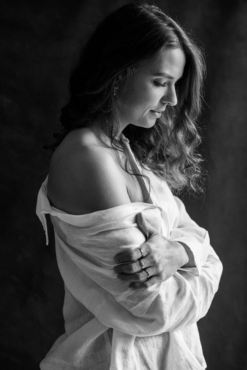 Vrouw in witte blouse met blote schouder, armen om zichzelf heen, gefotografeerd met eerlijke portretfotografie in Breda