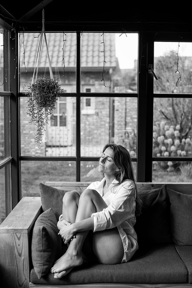 Zwart-wit portret van een vrouw zittend op een bank met opgetrokken knieën, gefotografeerd in een tuinkamer door een vrouwelijke fotograaf in Breda.