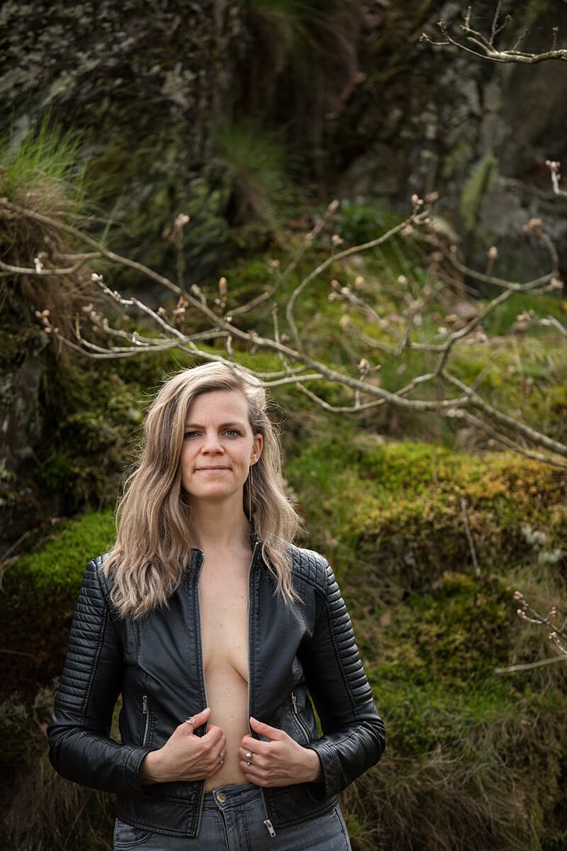 Zelfverzekerd portret van de vrouwelijke fotograaf in de Ardennen, poserend in een spijkerbroek en leren jasje met een subtiele glimlach.
