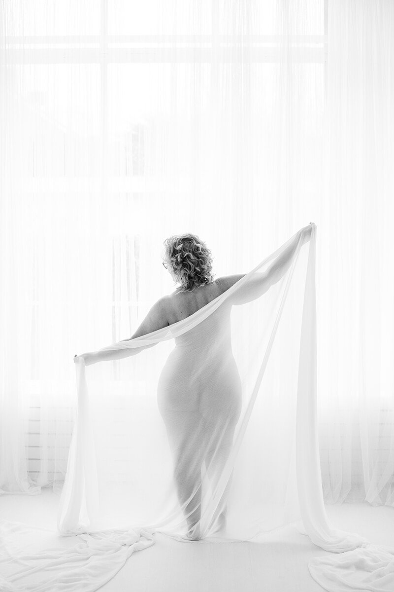 Boudoir fotografie Breda van plus size vrouw met chiffon doek in zacht tegenlicht bij witte gordijnen