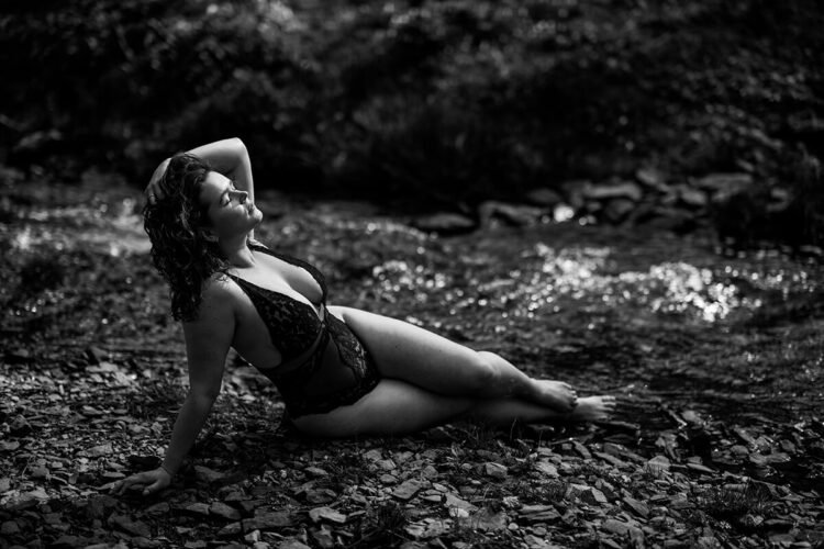 Sensuele boudoir foto in zwart-wit van een vrouw in zeemeermin-houding op een kiezelstrand bij een rivier in de Ardennen.