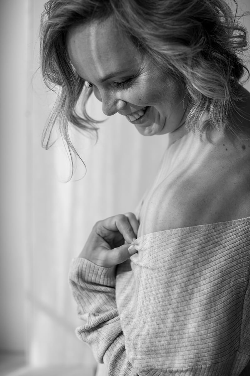Close-up van een lachende vrouw die over haar schouder kijkt terwijl ze haar trui vasthoudt, in zwart-wit.